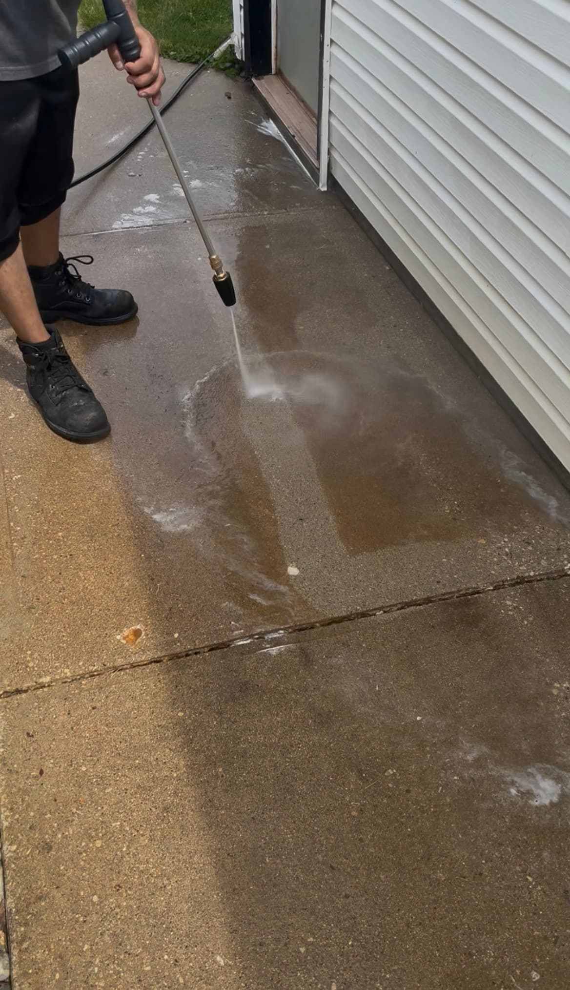 Person in black boots pressure washing a dirty concrete patio next to white house siding.
